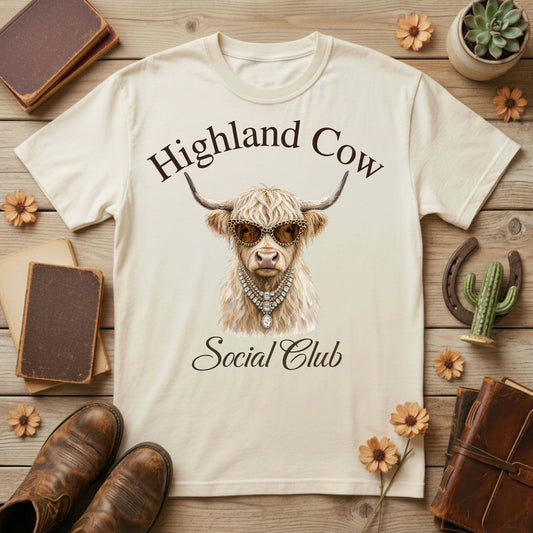 A cream-colored graphic t-shirt featuring a detailed illustration of a Scottish Highland cow wearing sunglasses and a necklace, with the text 'Highland Cow Social Club' printed around it. The t-shirt is displayed in a styled flat-lay arrangement on a rustic wooden background surrounded by vintage books, boots, a cactus, and flowers.