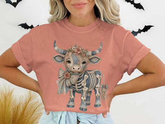 Skeleton highland cow graphic with flowers and bows on a peach t-shirt.