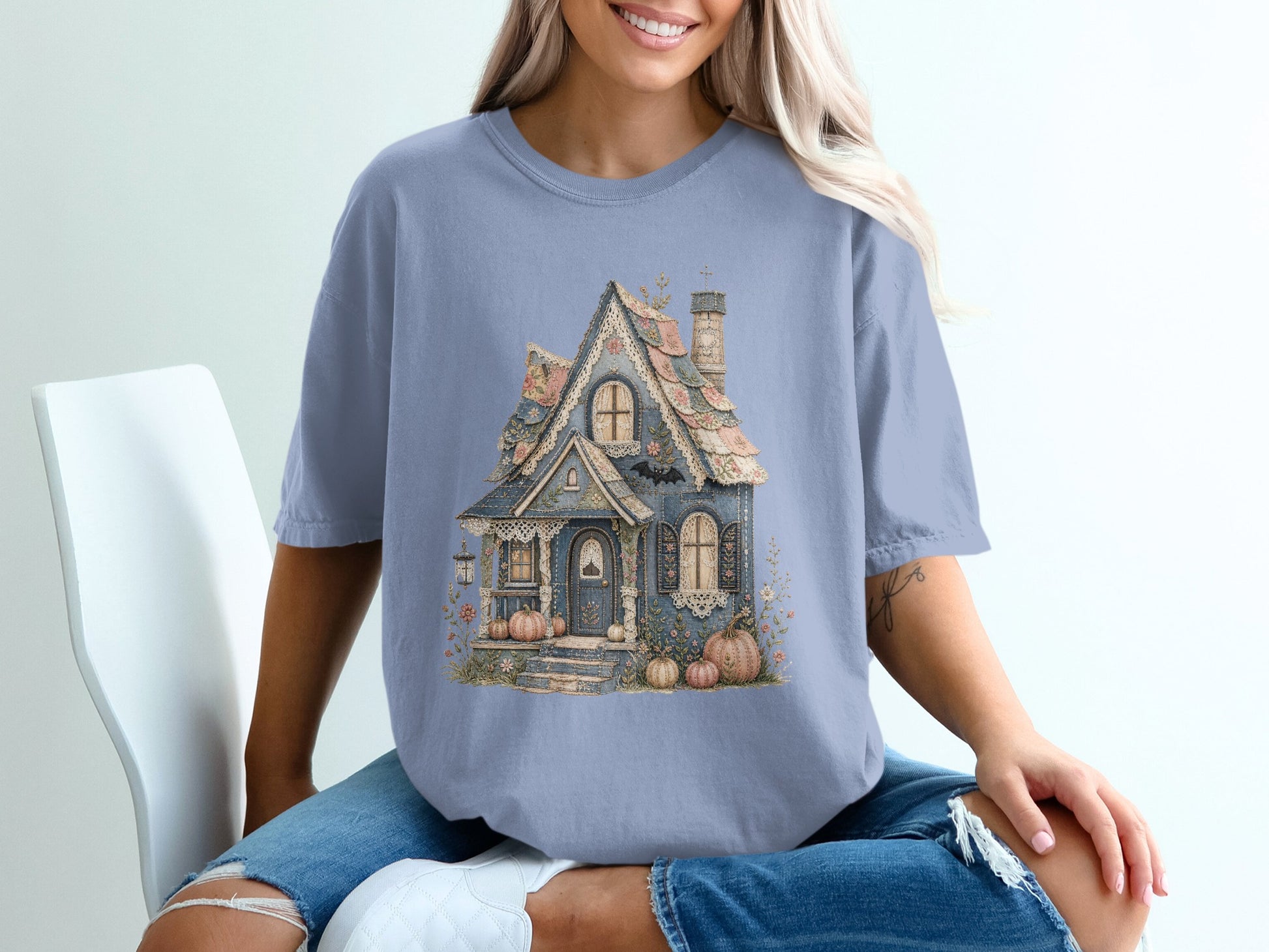 Blue t-shirt with a detailed illustration of a whimsical, pastel-colored cottage with pumpkins and a bat.