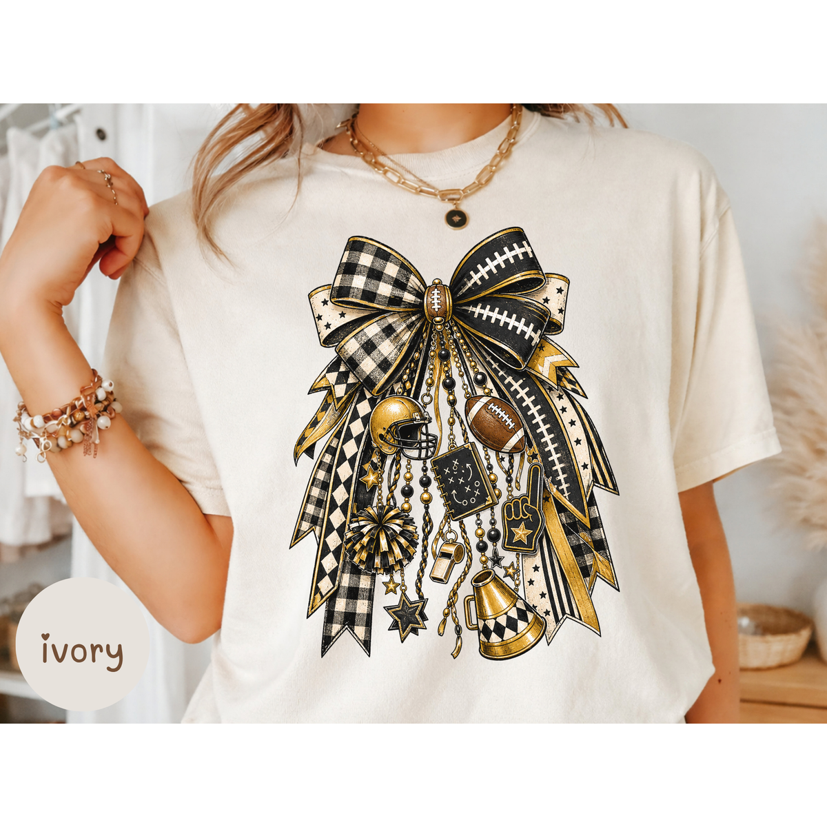 A person wearing an ivory short-sleeve T-shirt with a large black, white, and gold ribbon-and-bow graphic on the chest, styled with gold jewelry in an indoor setting. A small label in the lower left reads 'ivory'.